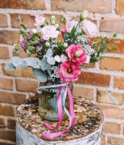 Colorful flowers. The bouquet in a decorative glass jar stands on the trunk of a tree. Brick wall in the background.